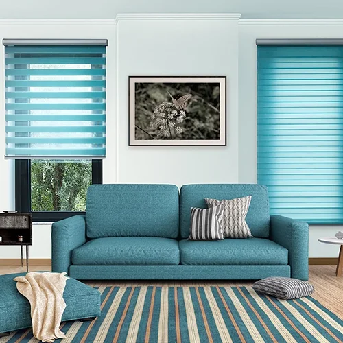 A modern living room with teal sofa, striped rug, JLS blinds, and a butterfly photo. Cushions are on the floor and sofa.