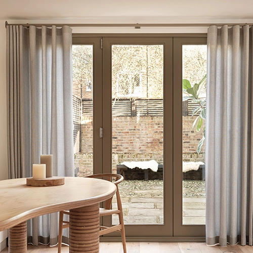 A modern room with floor-to-ceiling glass doors leading to a brick patio and fenced garden. Light grey made-to-measure curtains frame the doors, and a wooden table with candles and a chair are in the foreground.
