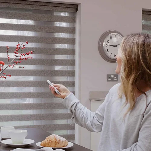 A woman uses a remote to adjust JLS blinds in a kitchen with cups, plates, and a croissant on the table.