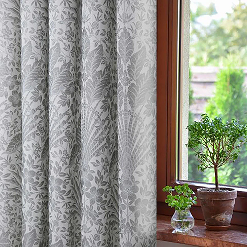 Grey floral-patterned Made to Measure Curtains partially cover a window with potted green plants on the windowsill and a view of trees and a fence outside.