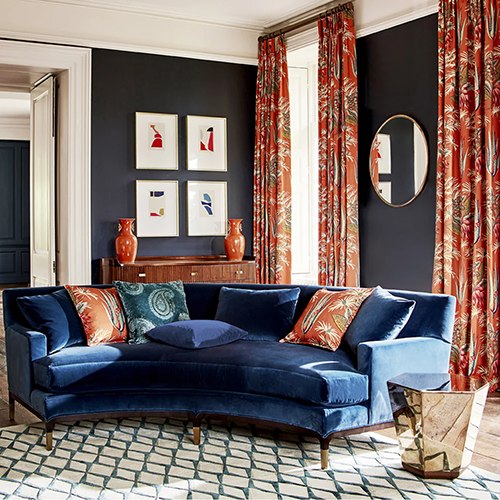 A stylish living room with a curved blue velvet sofa, patterned cushions, a geometric rug, made-to-measure curtains in red-orange floral print, framed modern art, two orange vases on a console, and a round mirror on a dark wall.
