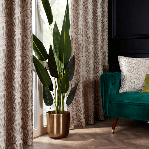 A tall leafy plant in a gold pot stands by a window adorned with Made to Measure Curtains, next to a green velvet sofa with a matching patterned cushion in a stylish lounge.