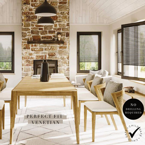 A modern dining room with a wooden table, six chairs, a stone fireplace, and large windows featuring Perfect Fit Venetian Blinds. The text reads Perfect Fit Venetian and No drilling required, with a child-safe certification icon.