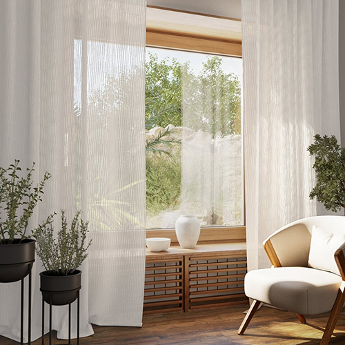 A cosy room with made-to-measure curtains framing a large window, letting in natural light. Potted plants, a modern chair with a cushion, and decorative vases on a wooden bench add charm. Lush greenery is visible outside.
