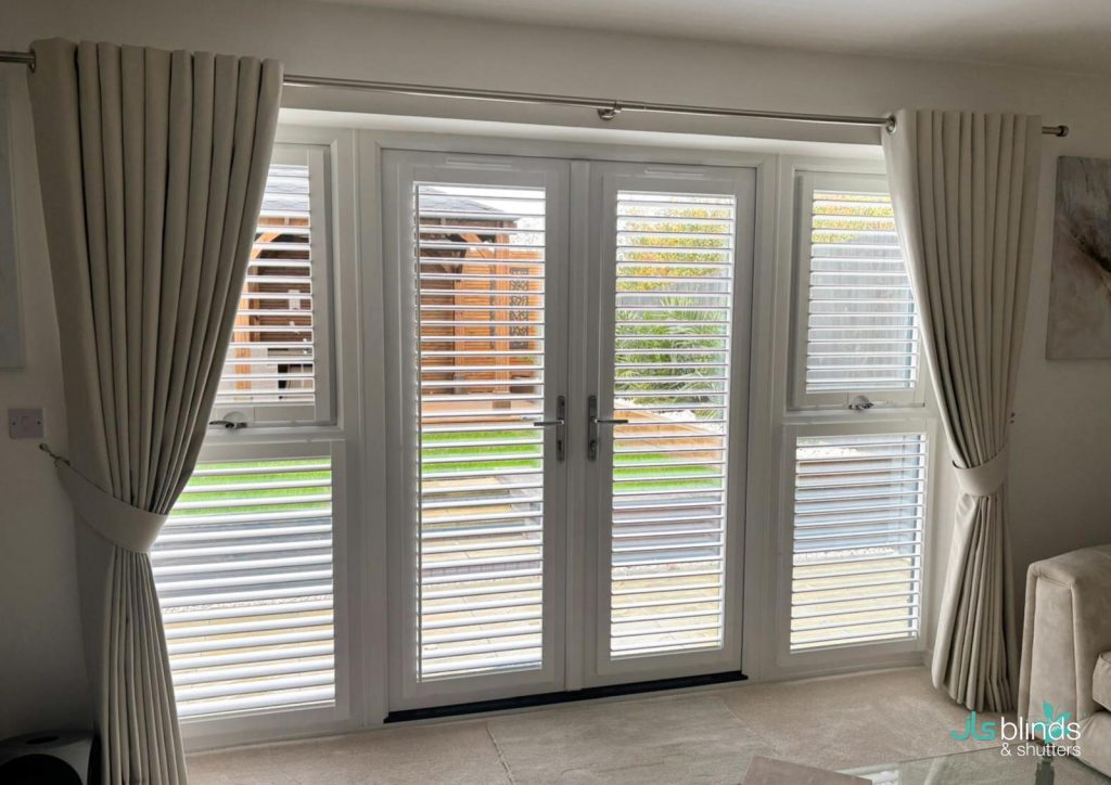 White plantation shutters cover glass patio doors with cream curtains in a bright living room; a garden and wooden structure are outside.