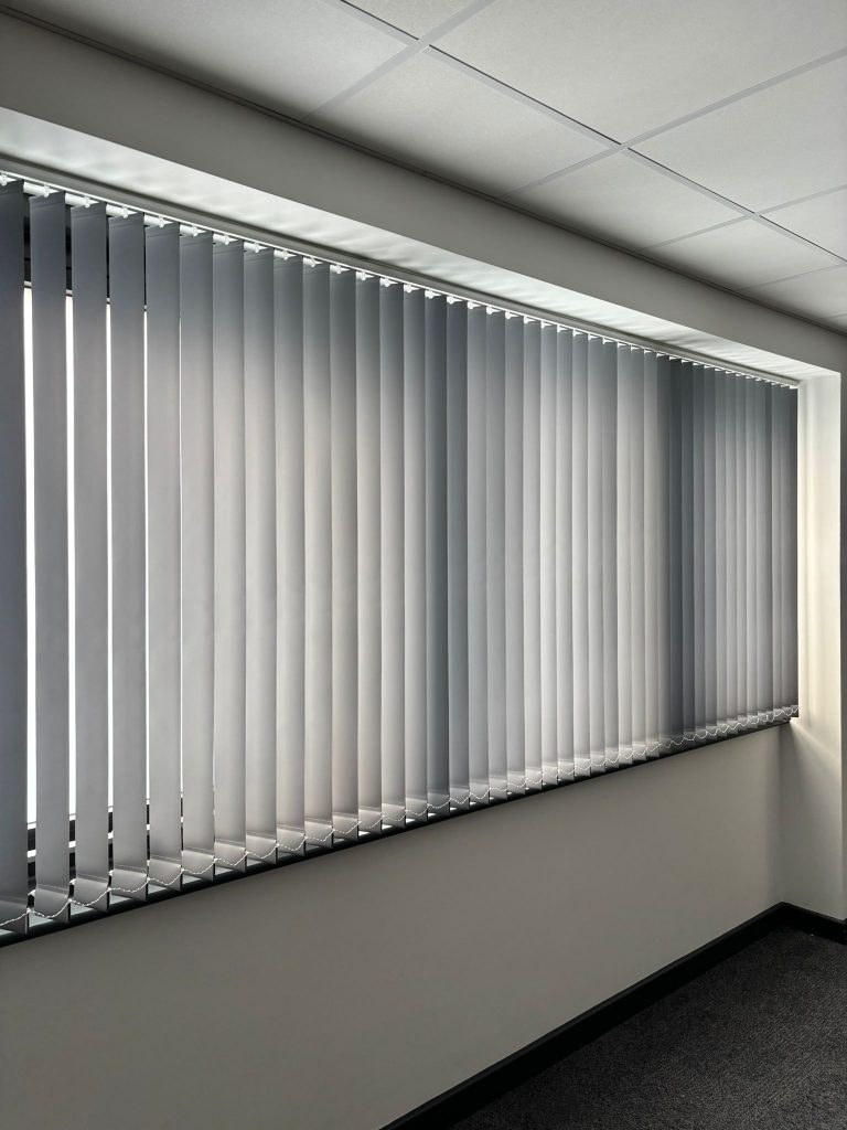 Vertical blinds partially closed over a large window in a modern, bright office space with white walls and ceiling tiles.