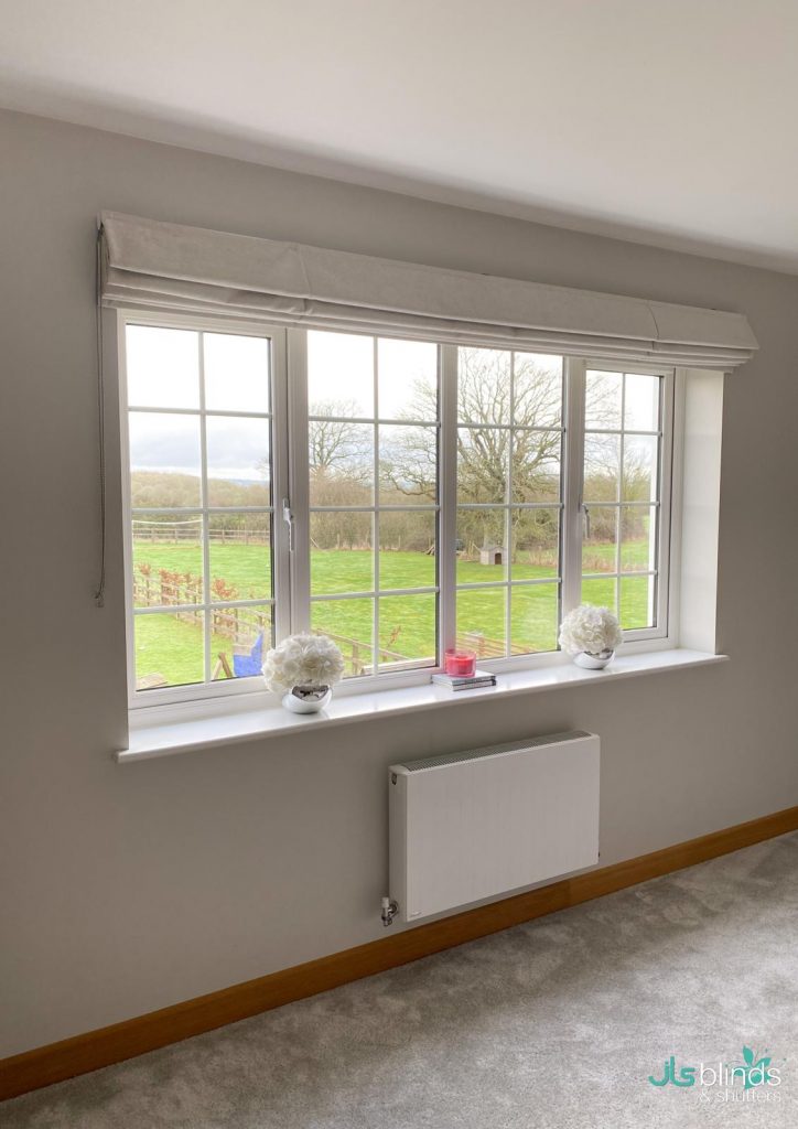 Large window with white Roman blind, two white flower arrangements, and a red candle on the sill, overlooking a green field.