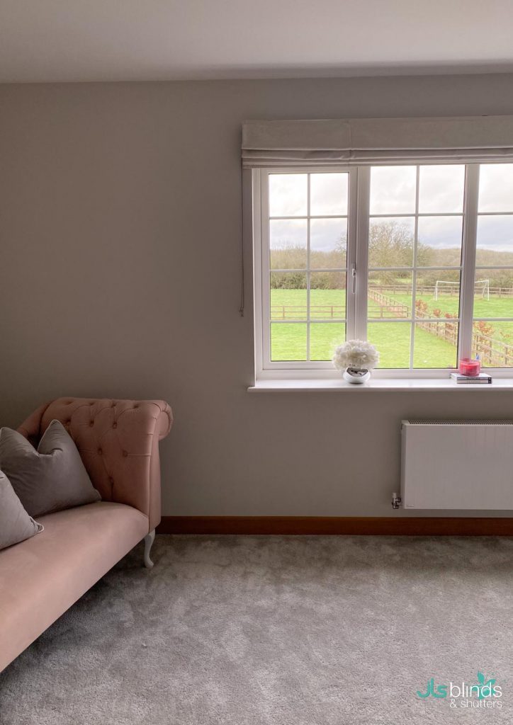 A cosy room with a pink sofa, grey cushions, and a window overlooking a green field and cloudy sky.