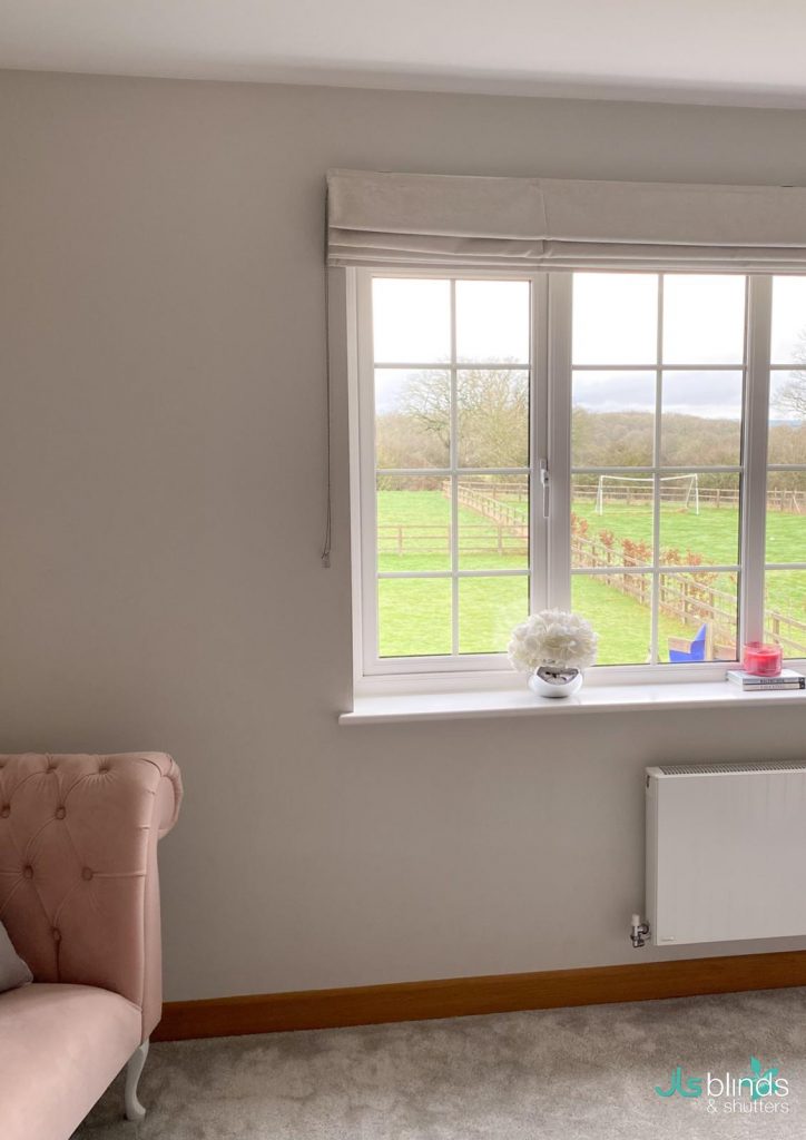 A large window with a roman blind overlooks a green field; a pink chair and a vase of white flowers sit nearby.