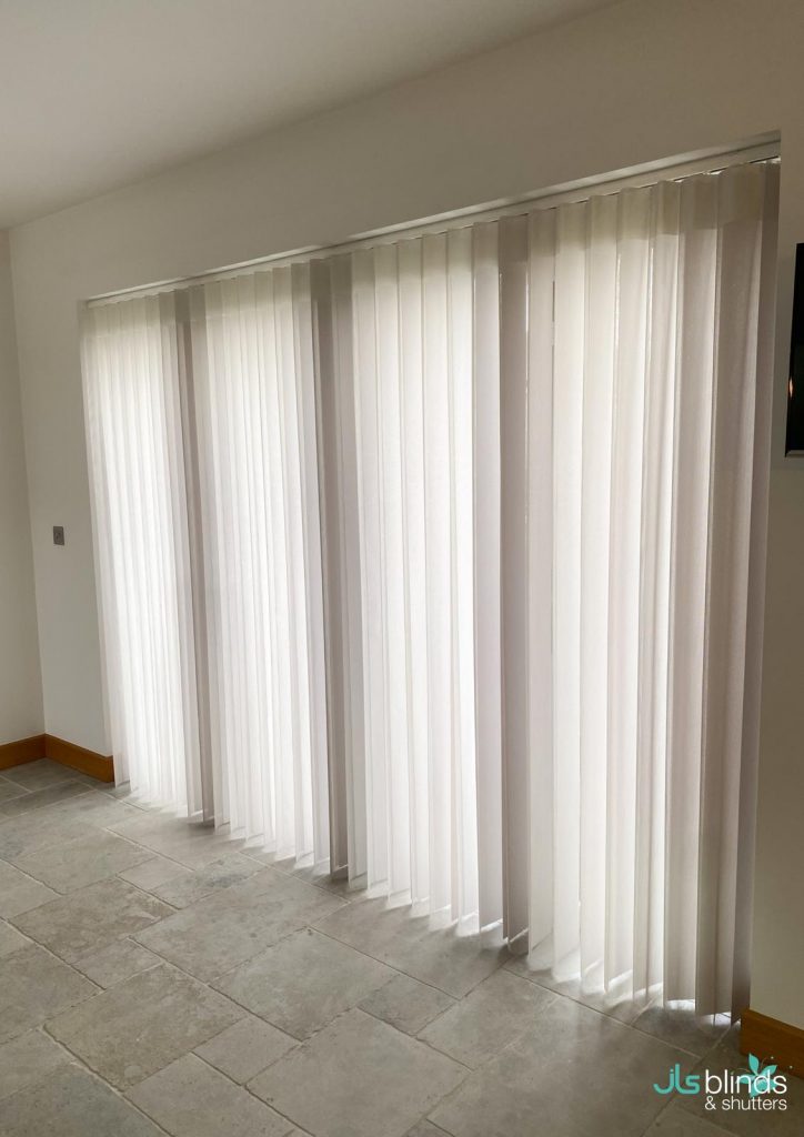 White vertical blinds cover large floor-to-ceiling windows in a bright room with light-coloured tile flooring.