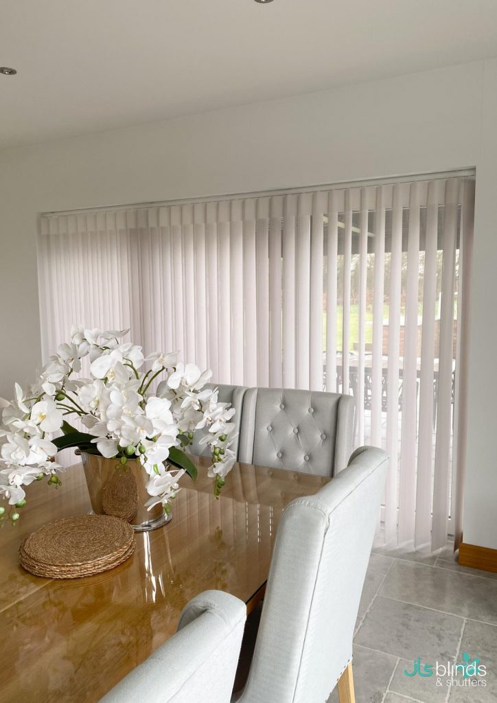 Dining area with a wooden table, grey chairs, a white orchid centrepiece, and vertical white blinds covering large windows.