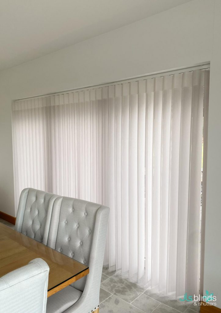 Vertical white blinds cover a large window beside a dining table and tufted upholstered chairs in a bright room.