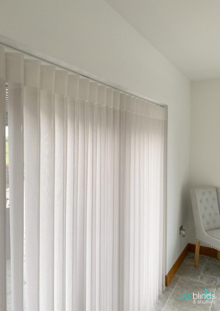 White vertical blinds cover a large window in a bright, modern room with a white accent chair in the corner.