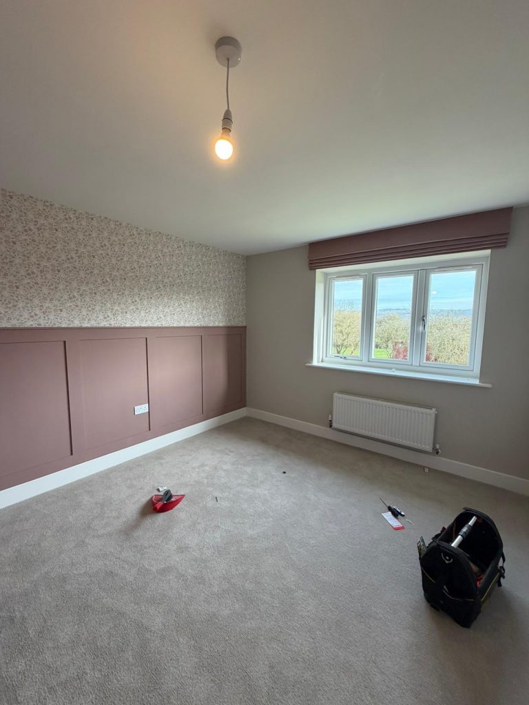 A mostly empty room with beige carpet, pink panelled walls, a single bulb light, and tools scattered on the floor near a window.