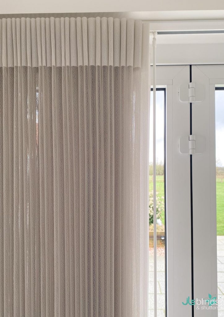 Beige pleated curtains next to glass patio doors, with a white vertical curtain pole and an outdoor view in the background.