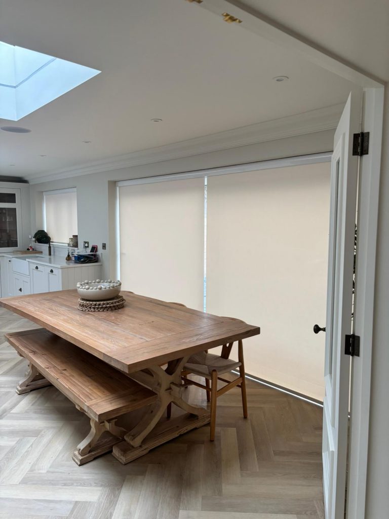 A wooden dining table with benches and chairs in a bright room with closed beige blinds and herringbone wood flooring.