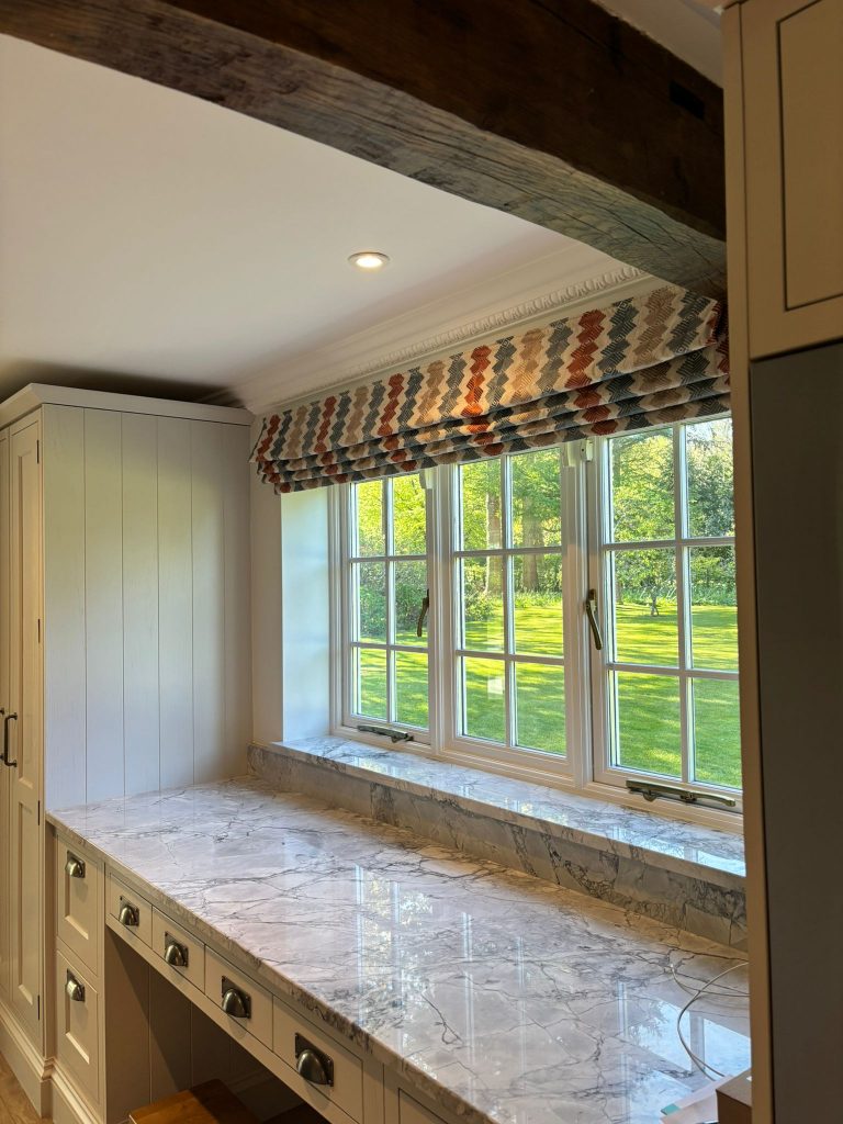 Sunlit kitchen with a marble worktop, large window, roman blind, and view of a green lawn and trees outside.