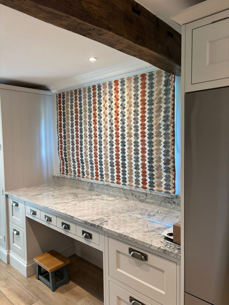 A kitchen workspace with a marble worktop, patterned window blind, white cupboards, and a small wooden step stool.