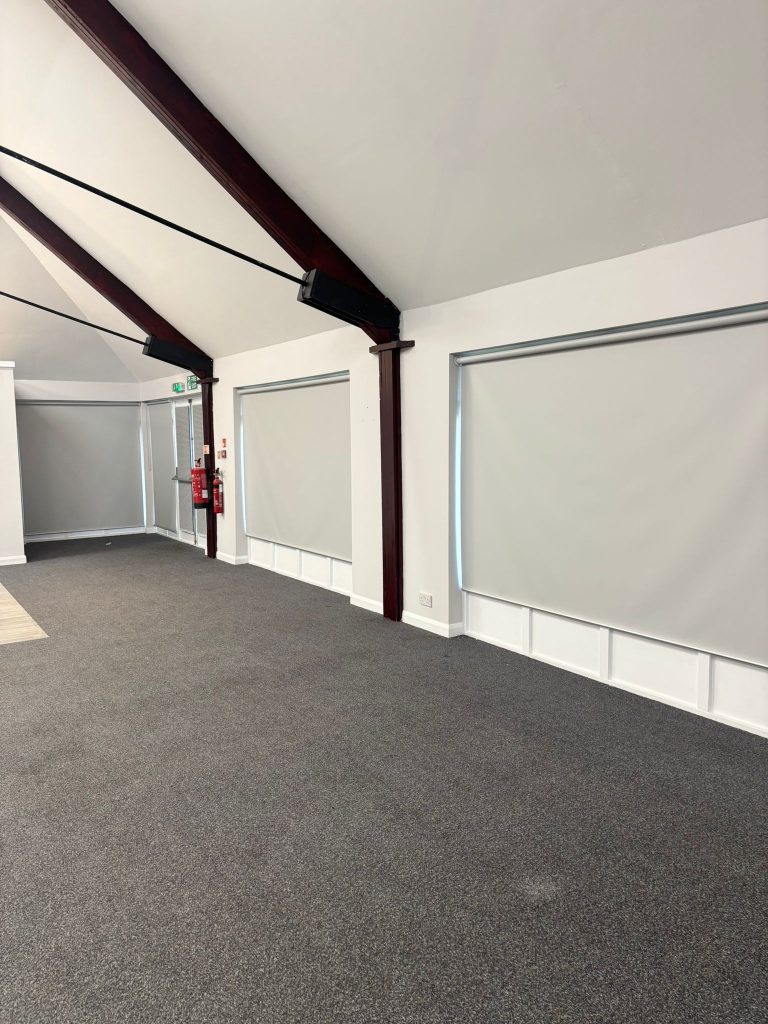 A spacious, empty room with grey carpet, white walls, covered windows, and exposed dark wooden beams on the ceiling.