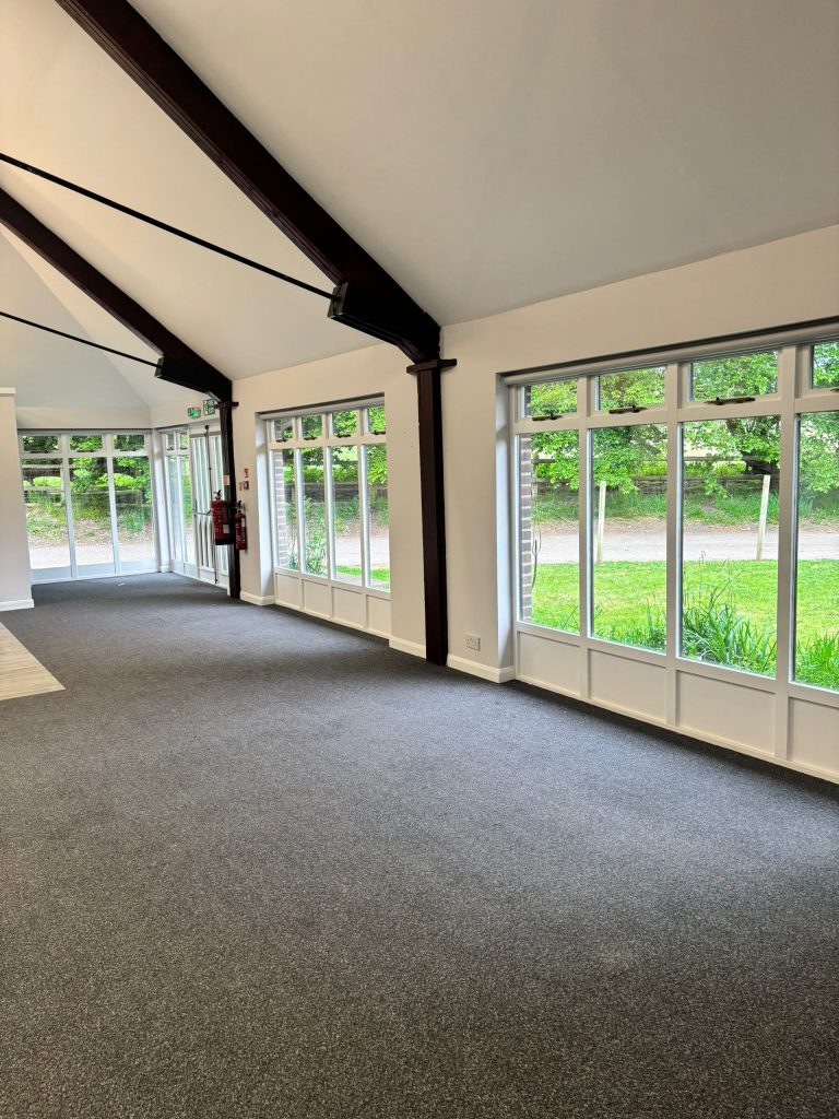 Spacious room with large windows, grey carpet, white walls, exposed dark beams, and scenic greenery outside.