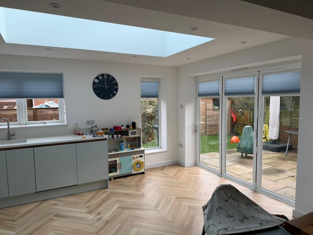 Bright modern kitchen and play area with wooden flooring, large windows, skylight, and toys by a garden patio.