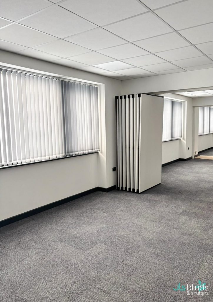 Modern office space with grey carpet, white walls, vertical blinds on the windows, and a folding partition wall partially open.