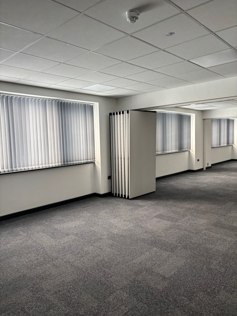 An empty office room with grey carpet, white walls, and large windows covered with vertical blinds.