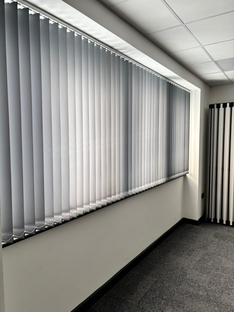 Large window with closed vertical blinds in a bright, modern room with white walls and grey carpet.