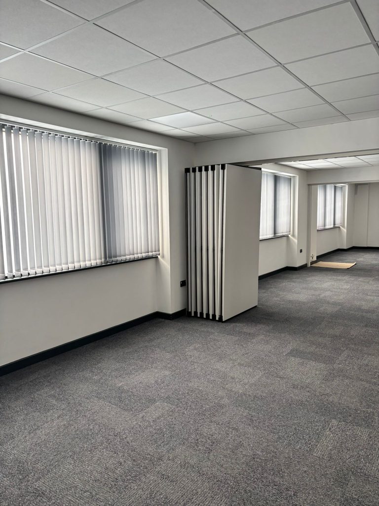 Empty office space with grey carpet, large windows with vertical blinds, and a folding partition wall.