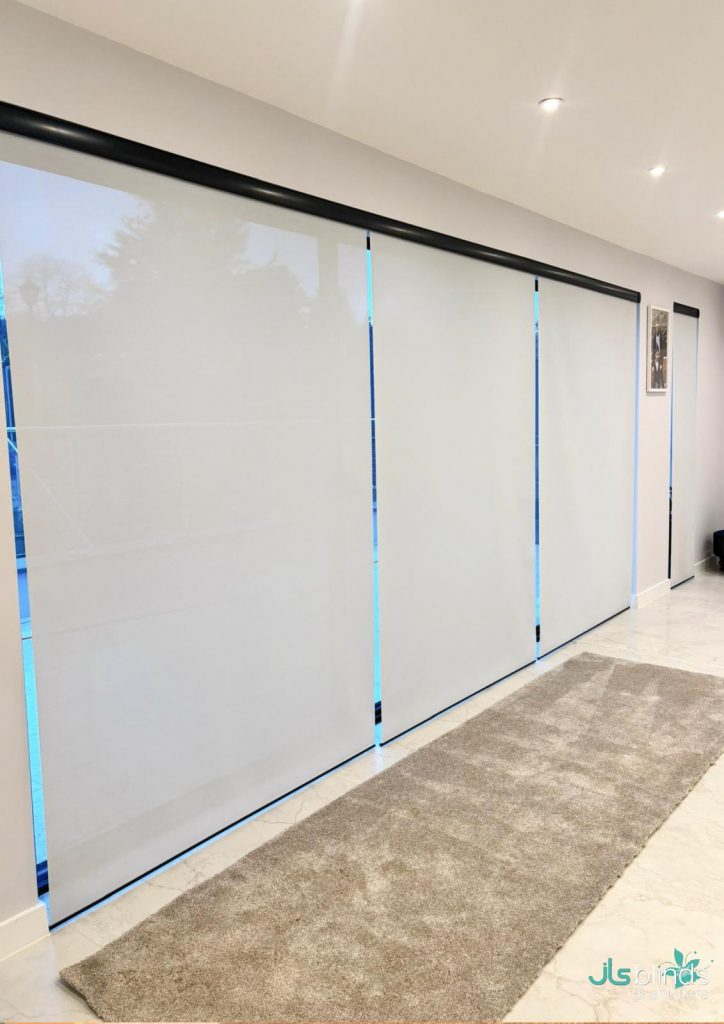 White roller blinds cover large windows in a modern room with a light rug on a tiled floor and a few art pieces on the wall.