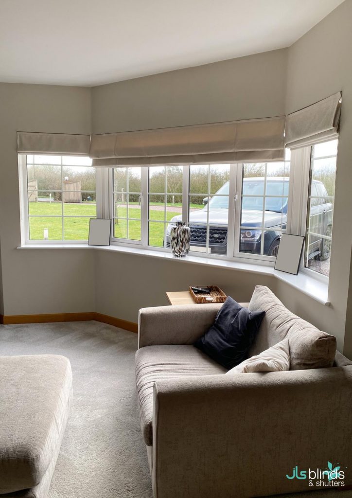 A cosy living room with a beige sofa by a large bay window, Roman blinds, and a view of a car and green lawn outside.