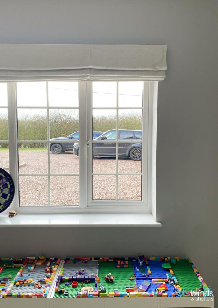 A window with white blinds looks out onto two parked cars; colourful building blocks are arranged on a table below the window.