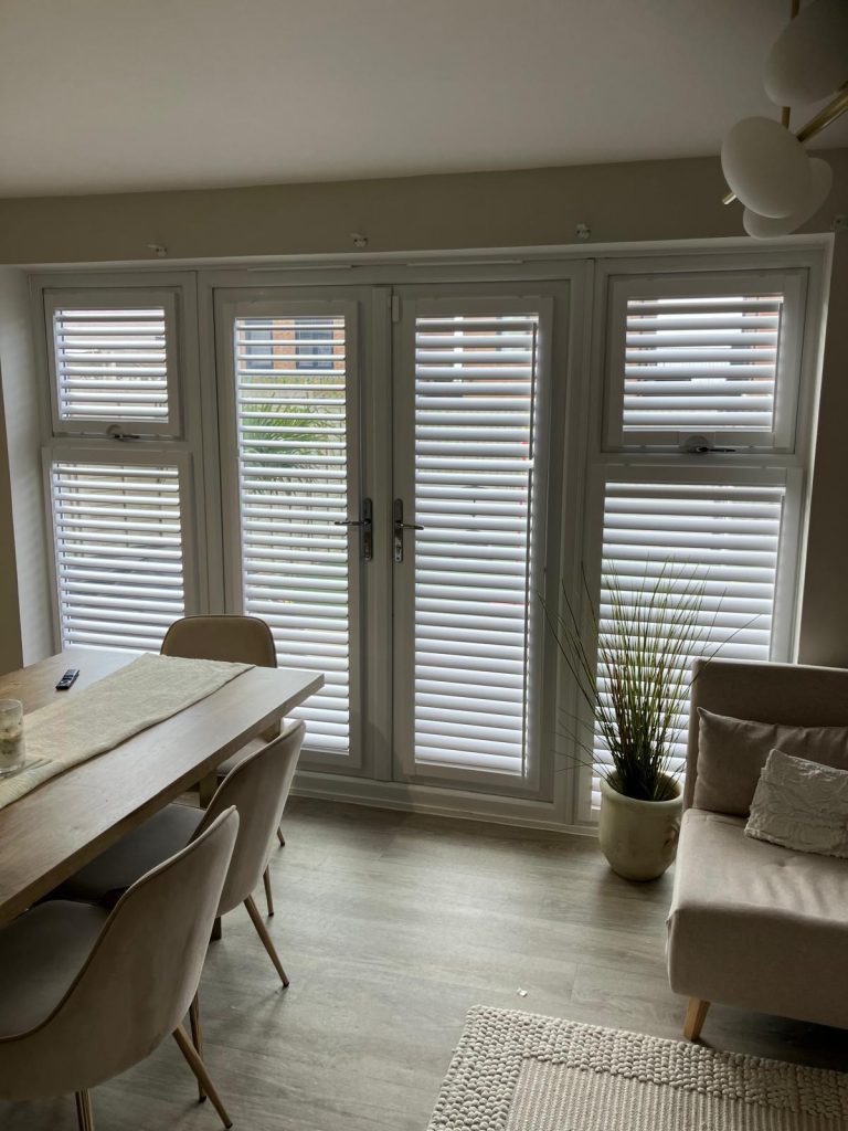 Dining area with a table and chairs beside patio doors and windows with white blinds, a potted plant, and a beige settee.