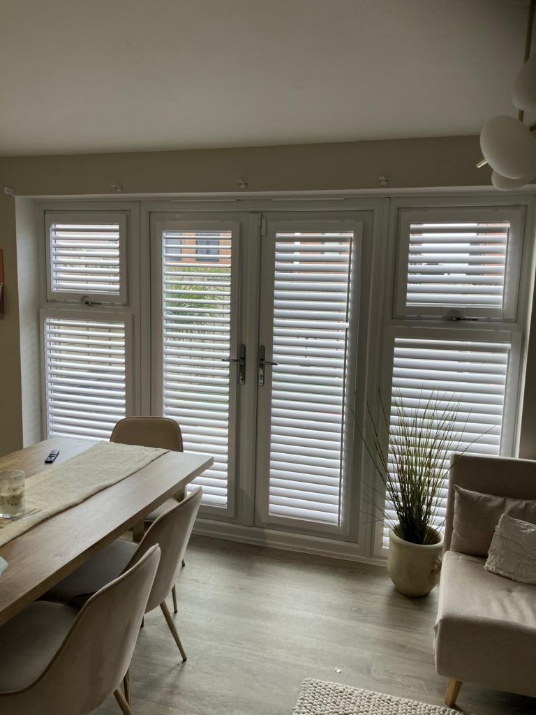 Bright room with a dining table and chairs, a small sofa, a potted plant, and large windows with white horizontal blinds.