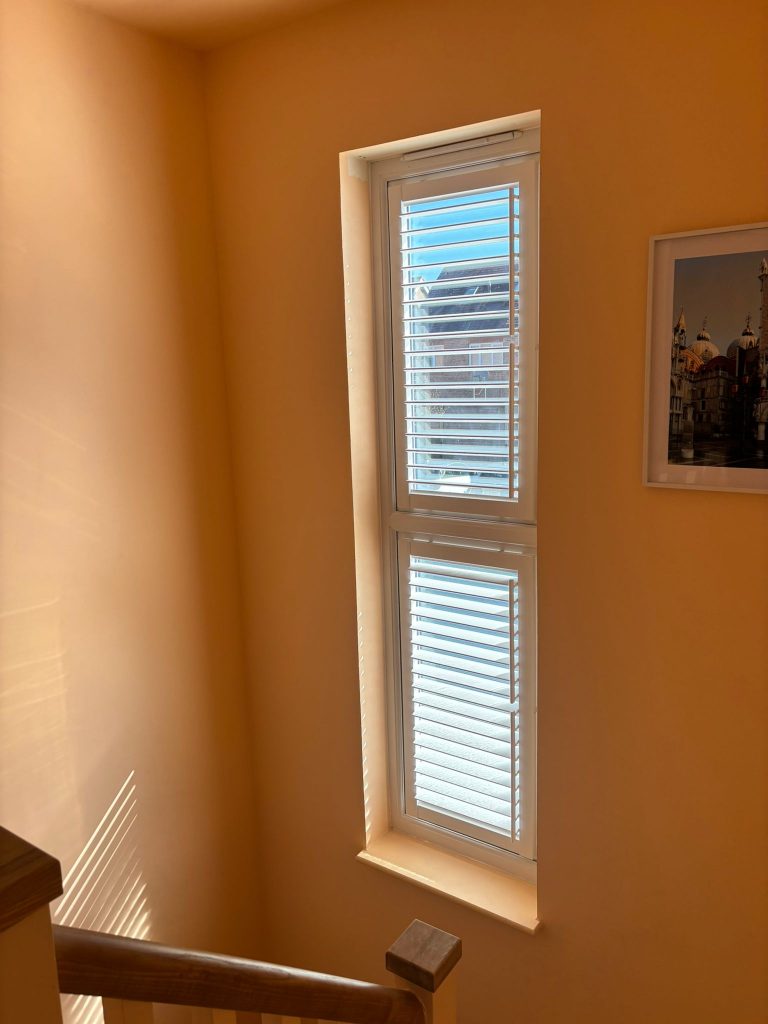 Sunlit stairway with wooden bannister, a tall window with white blinds, and a framed picture on a yellow wall.
