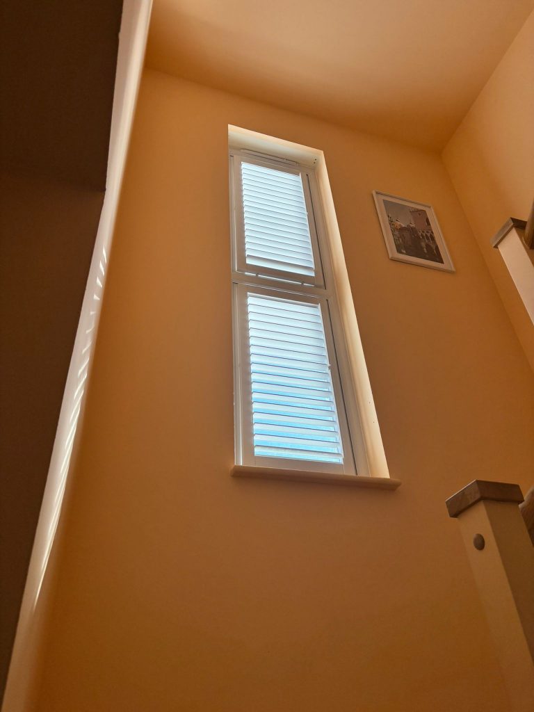 A tall window with closed blinds lets sunlight into a stairwell with peach-coloured walls and a framed picture hanging beside the window.