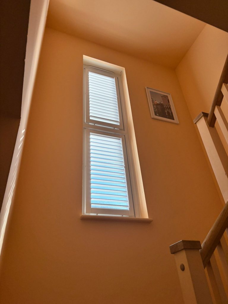 Tall window with closed blinds lets in light on a peach-coloured stairwell; a framed picture hangs on the wall near the stairs.