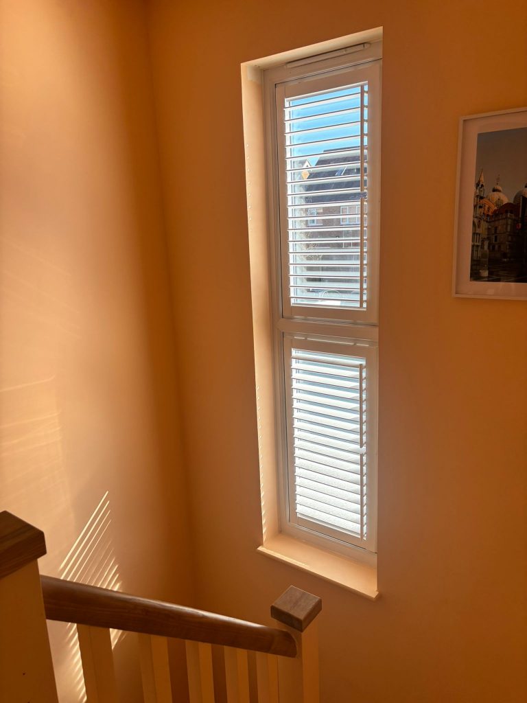 A sunlit stairwell with wooden bannister, tall window with white blinds, and a framed picture on a peach-coloured wall.