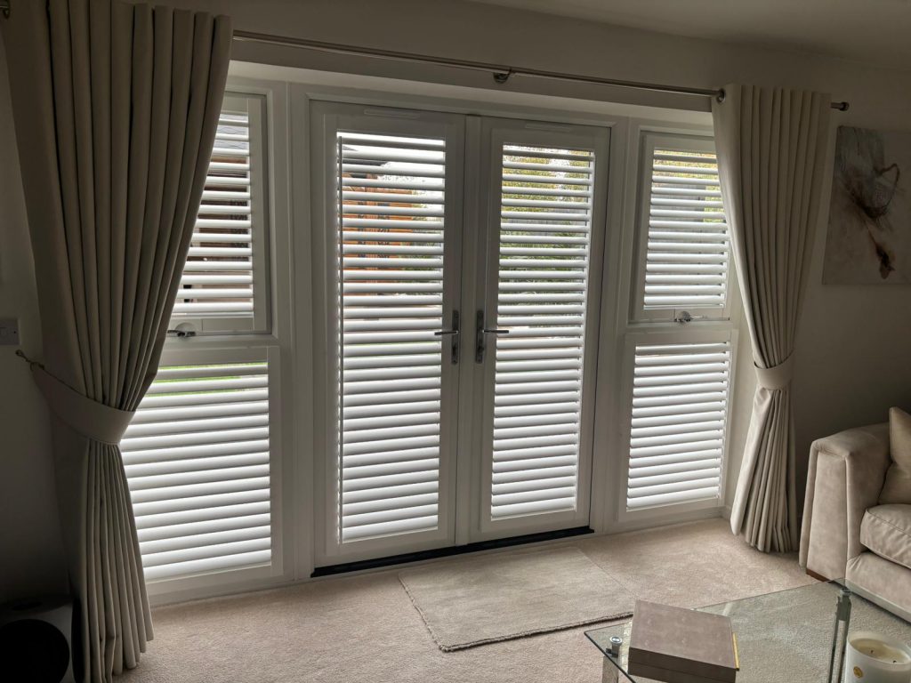 White plantation shutters and cream curtains cover large glass doors and windows in a bright, modern lounge.