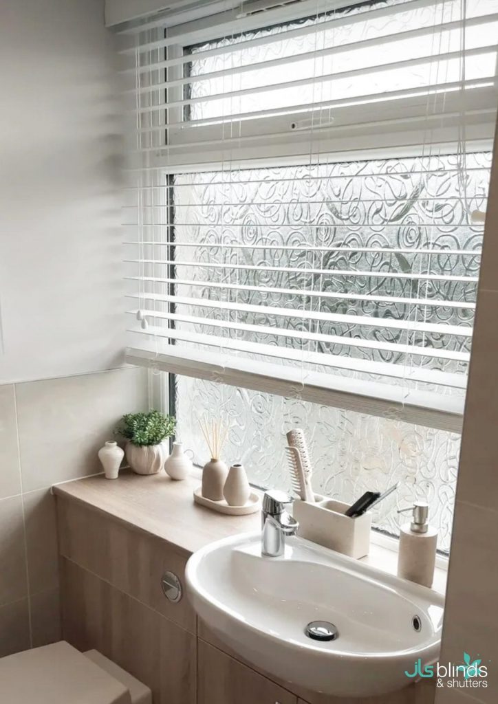 A bathroom with a sink, a shelf with decor items, and a window with white blinds and patterned privacy film.