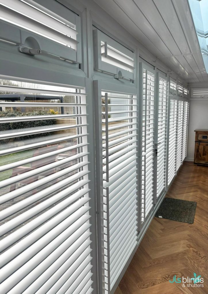 White plantation shutters cover large windows in a bright room with wooden flooring; outdoor scenery is visible through the slats.