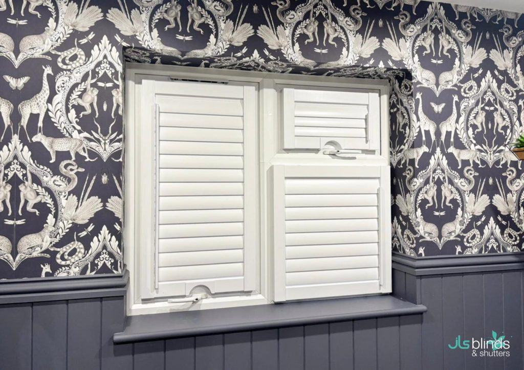 White shutter blinds on a window, set against dark patterned wallpaper with animal and floral motifs, and grey panelling below.