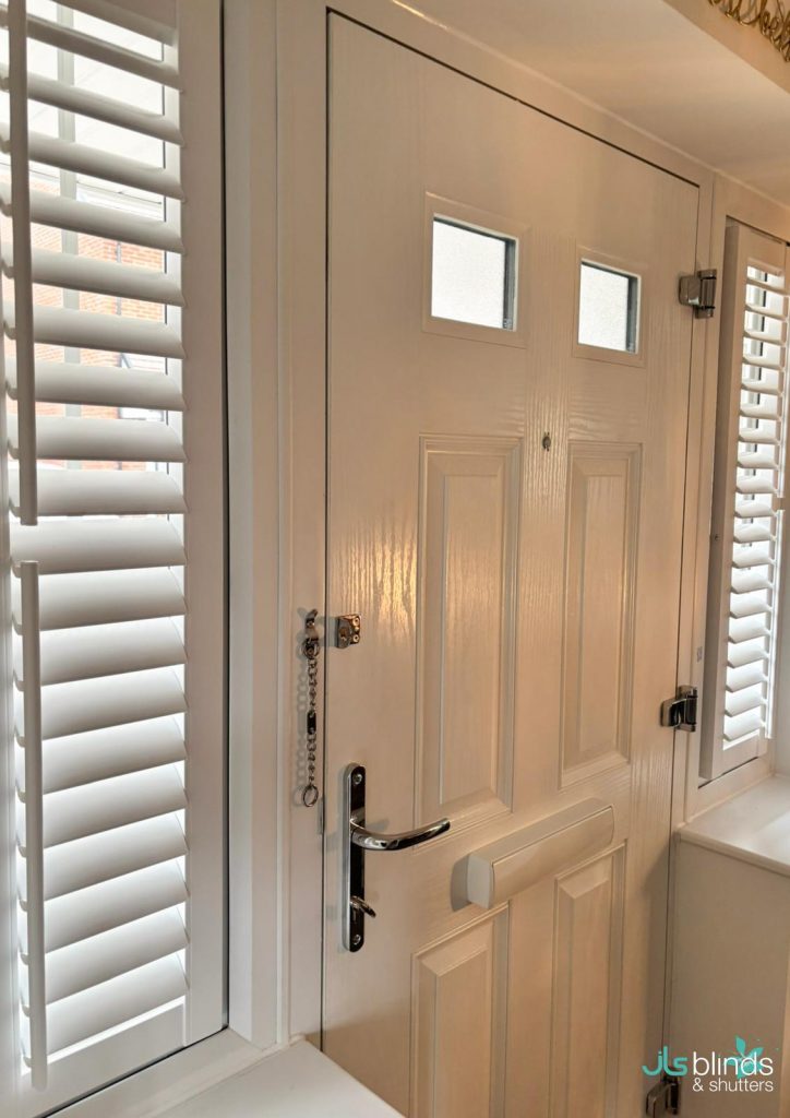 A white front door with a letter box, chrome handle, and two small windows, next to white plantation shutters on a window.