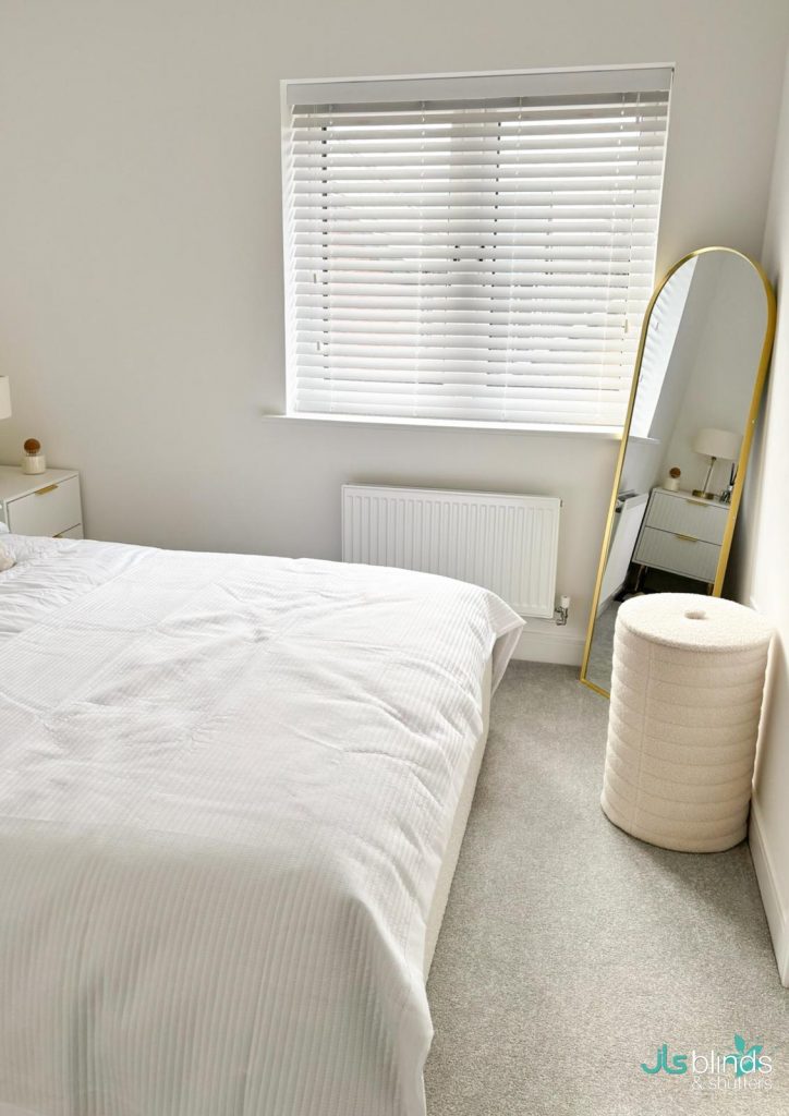 Minimalist bedroom with a white bed, white blinds on the window, a tall oval mirror, and a round beige pouffe on a grey carpet.