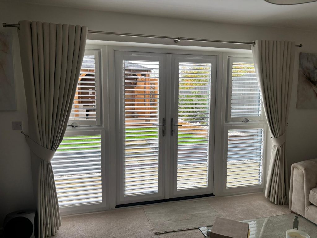 White French doors with shutters and cream curtains open to a garden with grass, a wooden pergola, and a paved patio outside.