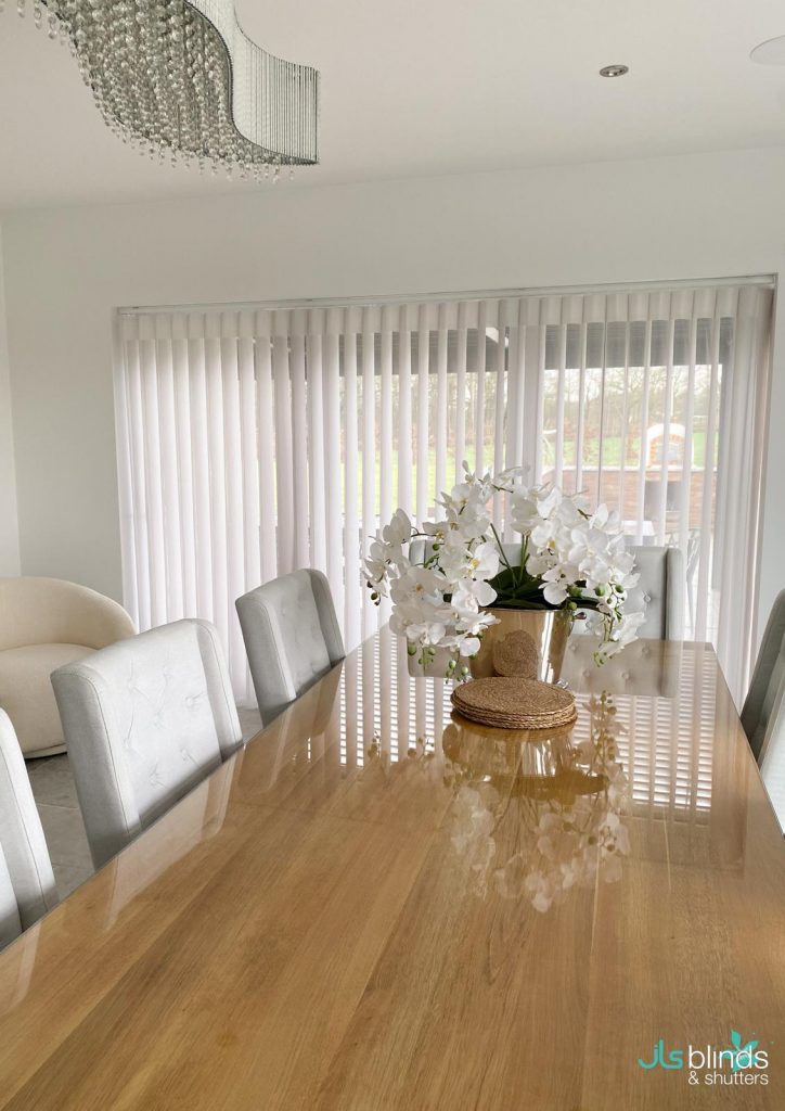 Modern dining room with a wooden table, white chairs, a vase of white flowers, and vertical blinds covering large windows.
