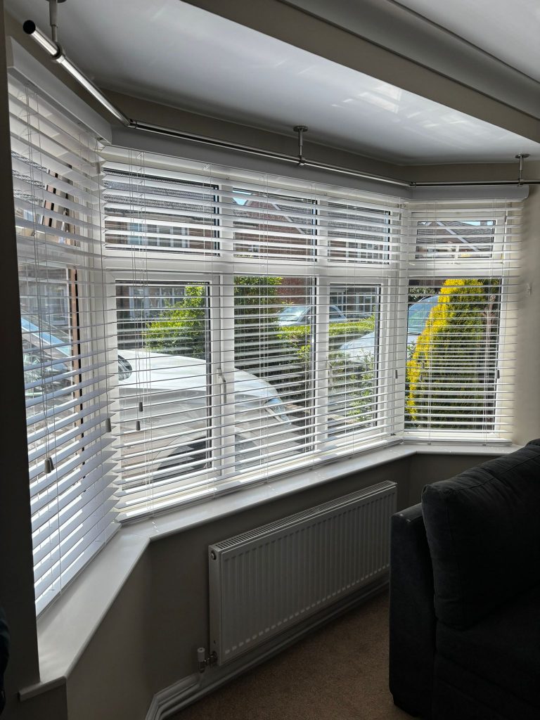 A sunlit bay window with white blinds, overlooking parked cars and houses, beside a grey sofa and a wall radiator.