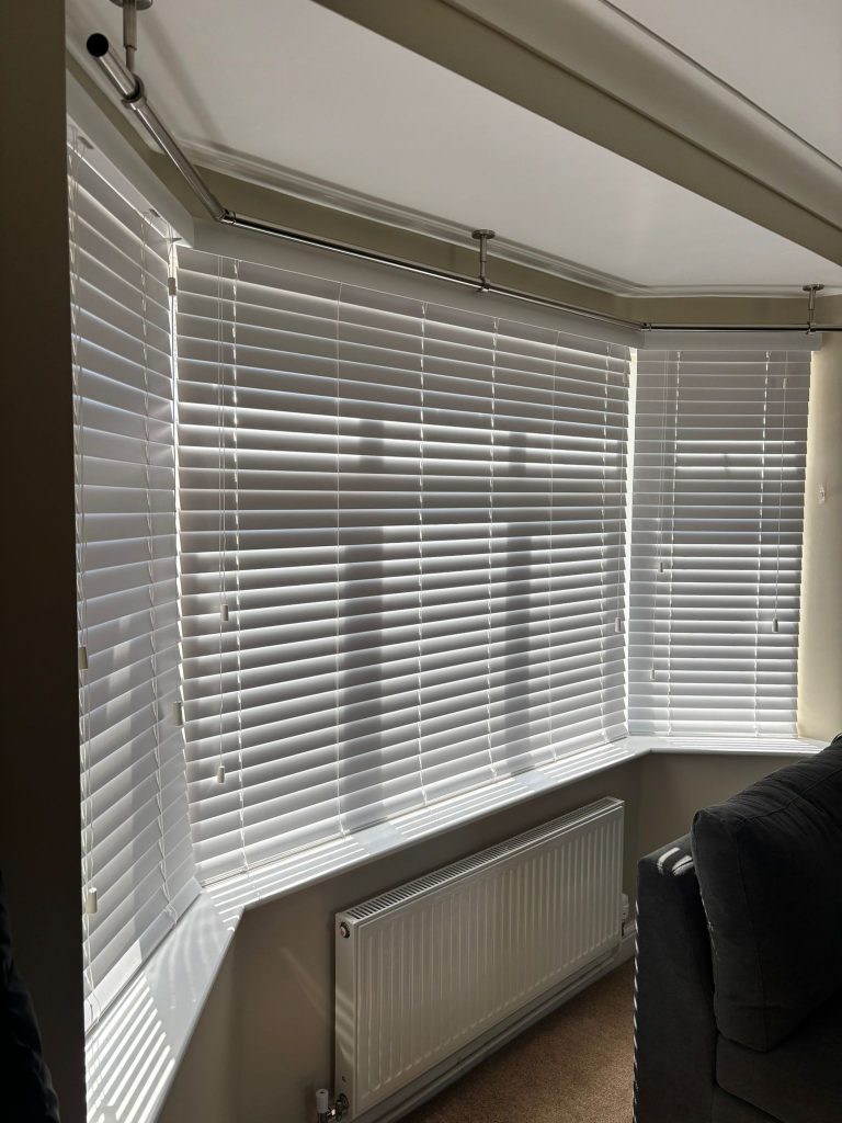 Sunlight casts the shadow of a cross on white window blinds in a bright living room with a radiator and part of a dark sofa visible.