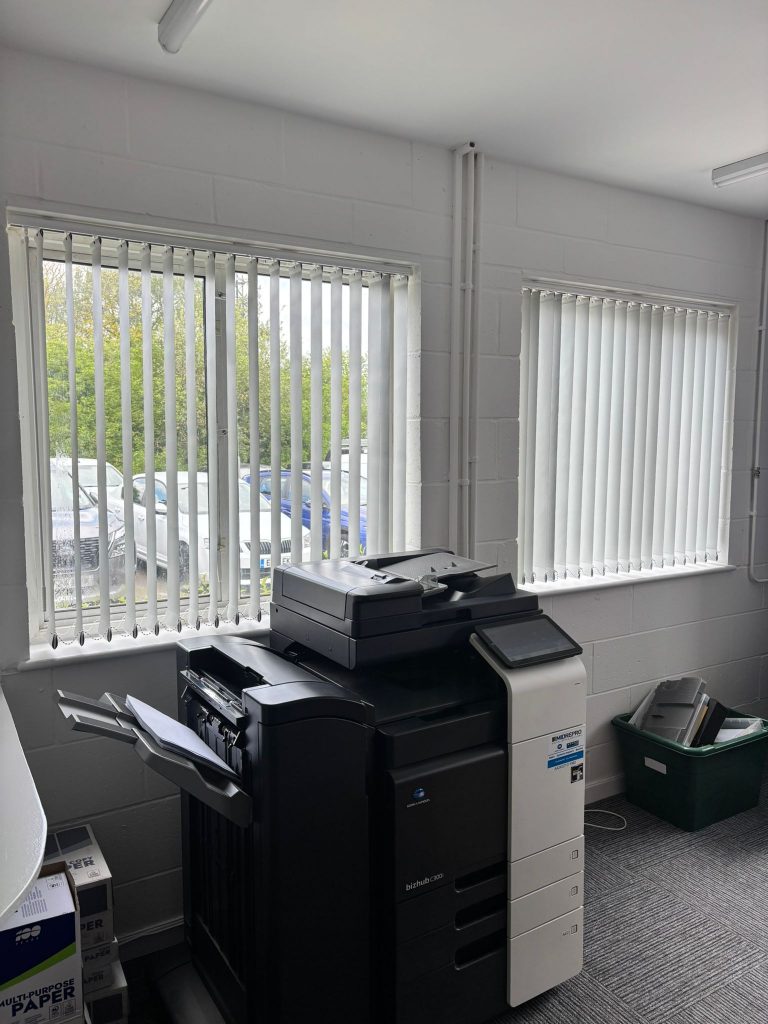A black office printer stands by two windows with vertical blinds in a bright room; a green recycling bin is on the floor nearby.