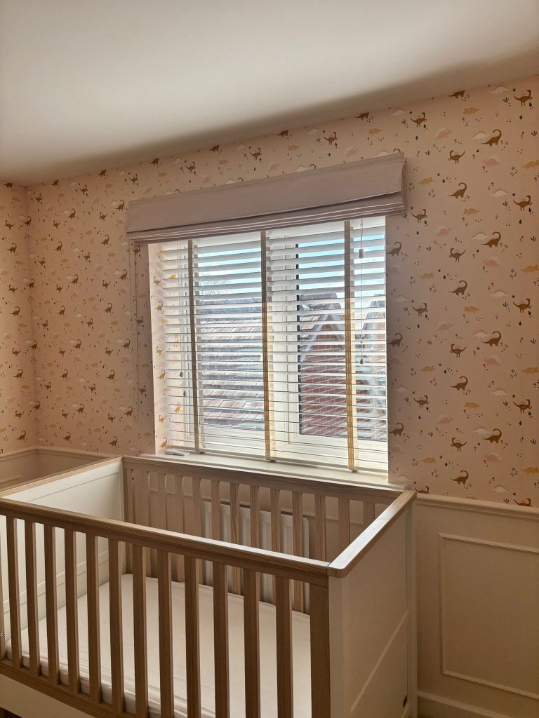 A white cot sits by a window with blinds in a nursery with animal-patterned wallpaper and soft natural light.